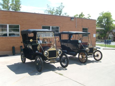 1912 Ford Model T - Two antique touring cars are parked side-by-side in front of a brick building on a sunny day.
