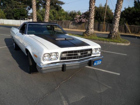 1973 Ford Ranchero - A white coupe utility with a black hood and vinyl roof is parked in a lot with palm trees.
