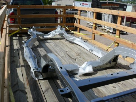 1976 Chevrolet Corvette - A car frame is secured with straps on a wooden trailer, with a pickup truck visible behind it.