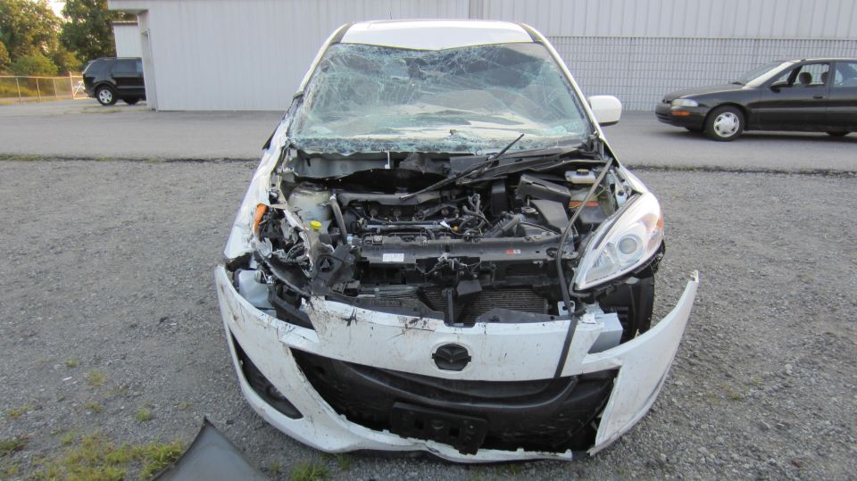 Front view of wrecked white 2012 Mazda Mazda 5 with hood open and windshield cracked. Another car is visible in the background