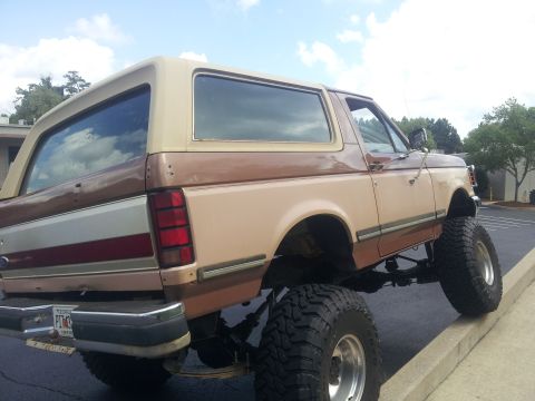 1988 Ford Bronco - This lifted Ford Bronco has a two-tone brown and tan paint scheme and is parked with its front tires on a concrete curb.