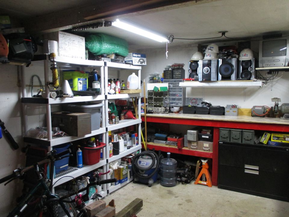 Garage workshop with shelves filled with supplies, tools, and storage bins. A workbench with equipment and a black filing cabinet are visible