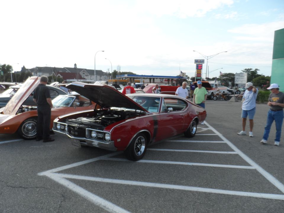 Red muscle car, possibly Oldsmobile Cutlass, parked at car show. Hood is open, showing engine. Another orange car is parked to the left