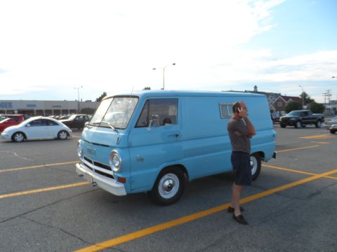 1971 Ford Ranchero - A man stands on his phone next to a light blue Dodge A100 van in a parking lot.