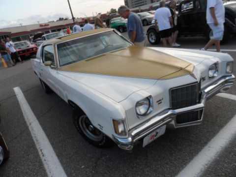 1971 Ford Ranchero - A white and gold 1973 Pontiac Grand Am coupe is parked at an outdoor event, with other vehicles and people in the background.