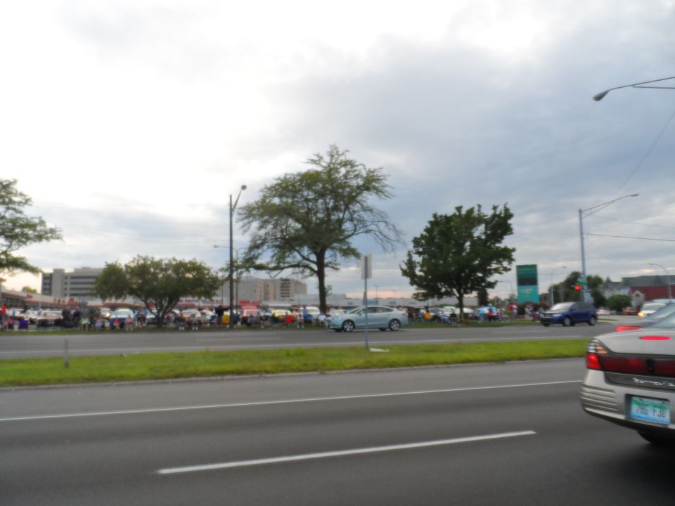 Cars lined up at a show with people gathered, a light blue sedan in the foreground