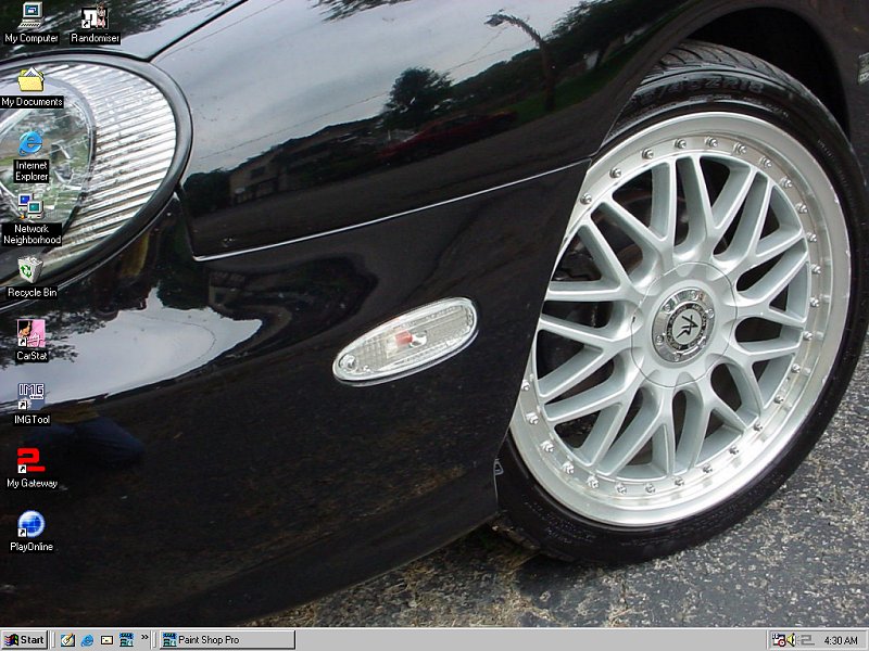 Close-up of a black car's front fender and wheel with aftermarket rims. Computer desktop icons are visible on the screen reflection