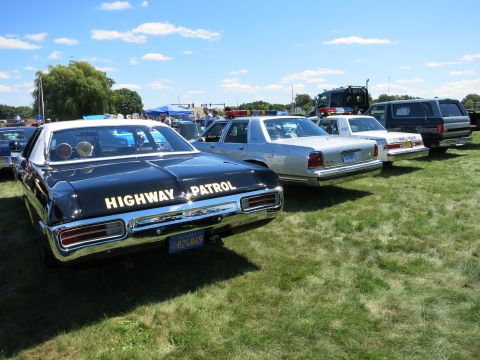 2001 Ford Crown Victoria - Several police cars, including a classic black and white model, are parked on a grassy field under a blue sky.