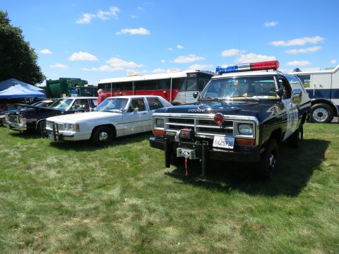 2001 Ford Crown Victoria - Several vehicles, including a police SUV and a white sedan, are parked on a grassy field under a blue sky.
