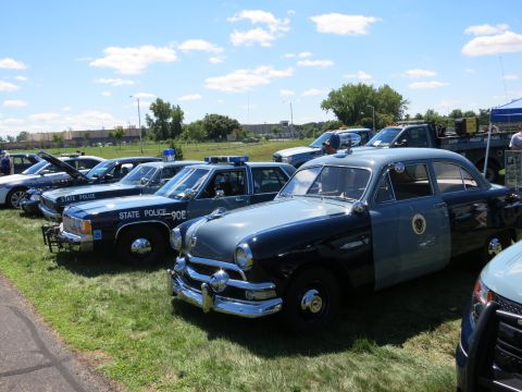 2001 Ford Crown Victoria - A line of police cars, including a vintage two-tone model, are parked on a grassy field under a partly cloudy sky.
