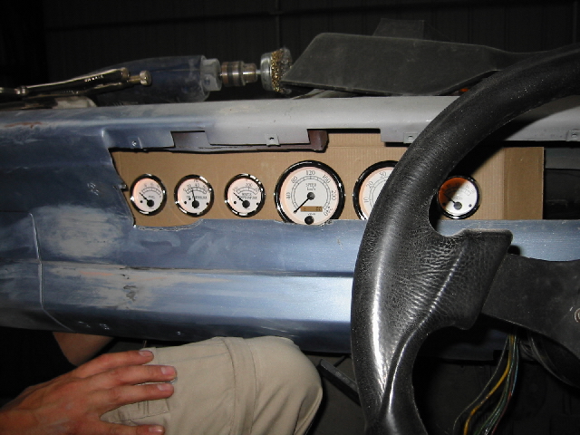 Aftermarket white-face gauges mocked into a cardboard dash panel on a bare-metal car