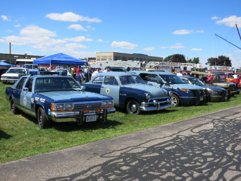 2001 Ford Crown Victoria - Several police cars, including a 1990s model and an older sedan, are lined up on a grassy field under a blue sky.