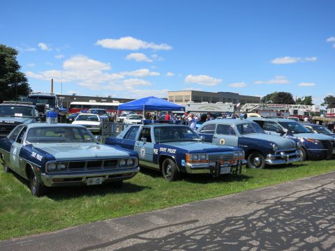 2001 Ford Crown Victoria - Several police cars are parked on a grassy field under a blue sky, with buildings and a fire truck in the background.