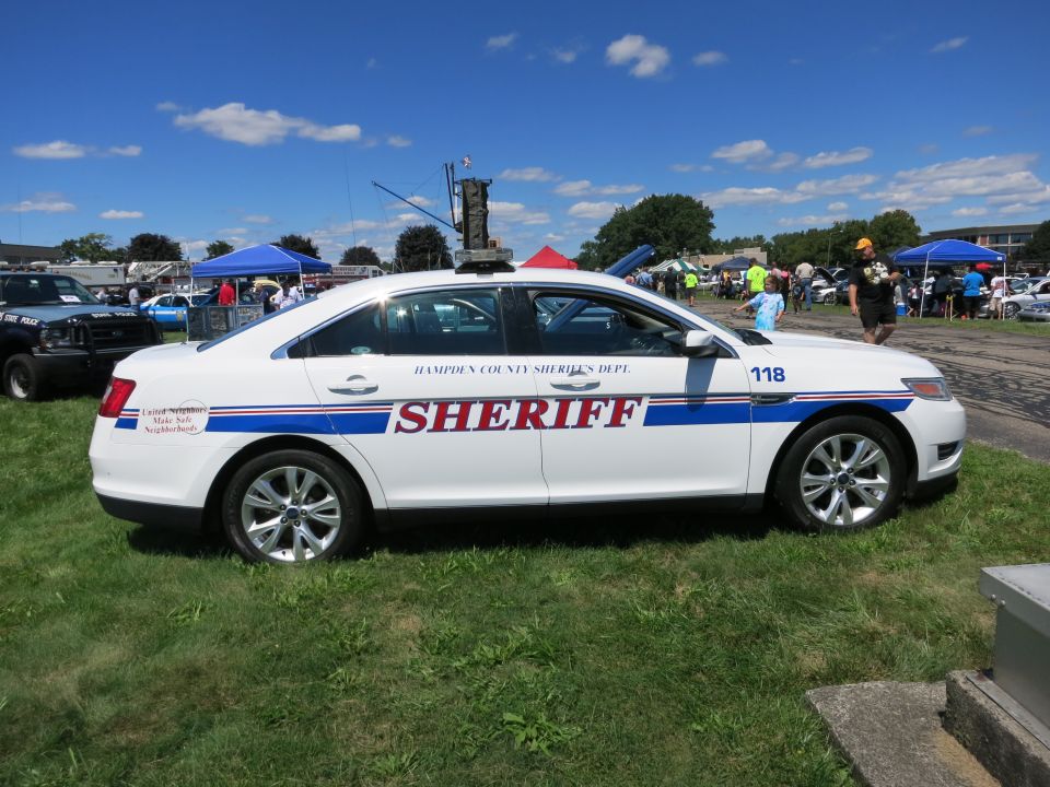 White sheriff's department sedan with 'SHERIFF' and 'HAMPDEN COUNTY SHERIFF'S DEPT.' text, parked on grass at an outdoor event