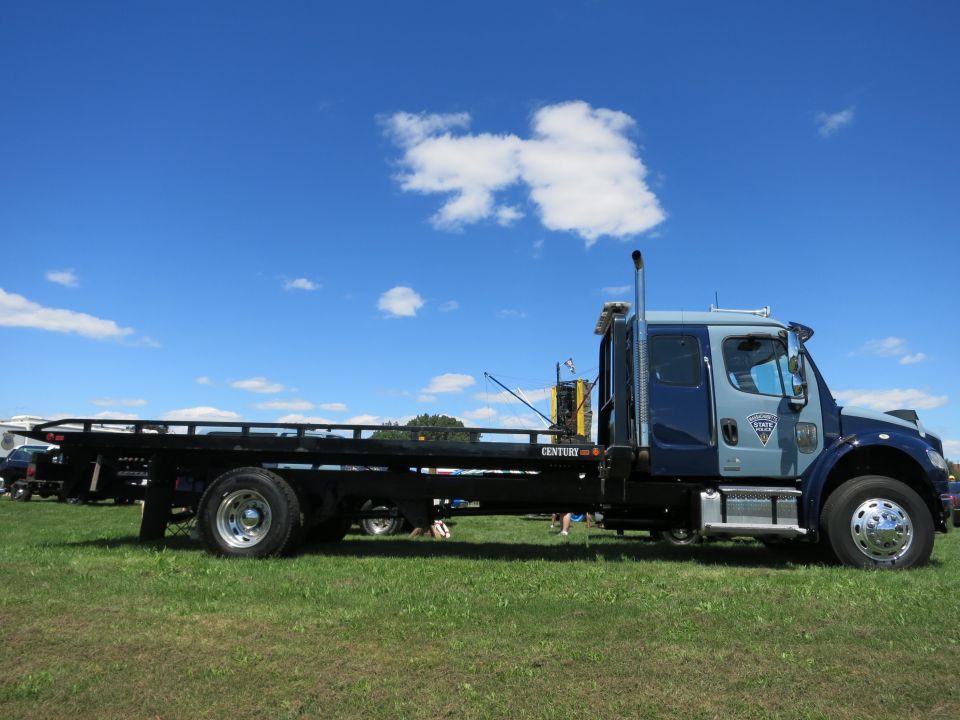 Blue and gray tow truck with a flatbed, parked on grass under a blue sky