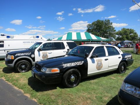 2001 Ford Crown Victoria - Two police vehicles, a sedan and an SUV, are parked on a grassy field under a clear blue sky.