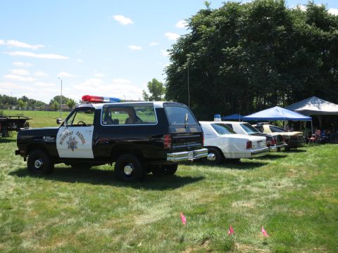 2001 Ford Crown Victoria - Several police vehicles, including a black and white SUV, are parked on a grassy field under a blue sky.