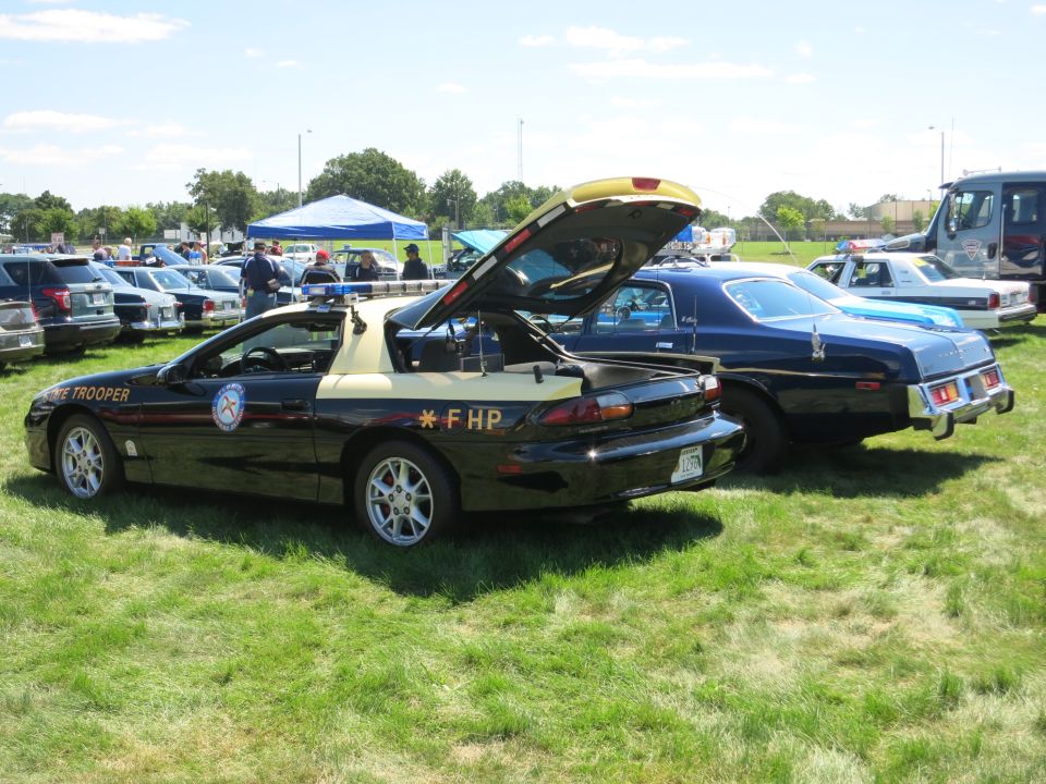 Black and tan police car with trunk open, parked on grass next to a blue sedan