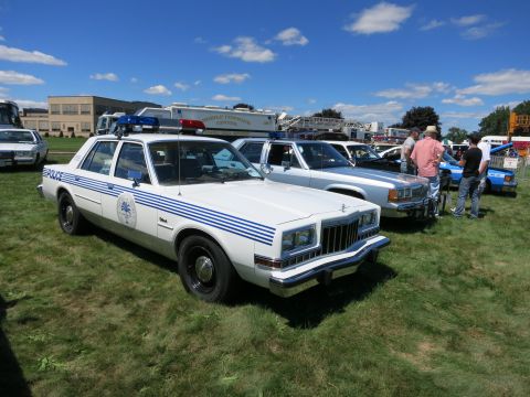 2001 Ford Crown Victoria - Multiple police cars are parked on a grassy field under a blue sky with clouds.