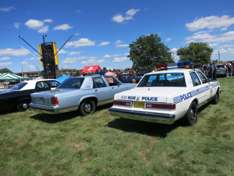 2001 Ford Crown Victoria - Two vintage police cruisers are displayed outdoors on a sunny day with a clear blue sky.