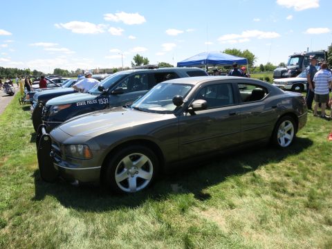2001 Ford Crown Victoria - A lineup of police vehicles, including a Dodge Charger and a Ford Explorer, are parked on a grassy field under a sunny sky.