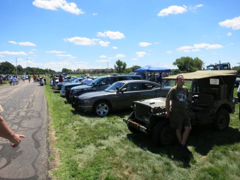 2001 Ford Crown Victoria - A military jeep and several police cars are parked in a line on a grassy field on a sunny day.