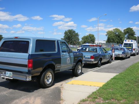 2001 Ford Crown Victoria - A line of police vehicles, including a Bronco and Crown Victorias, are parked along a curb under a blue sky with clouds.