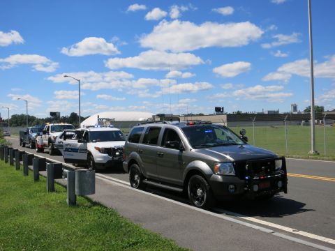 2001 Ford Crown Victoria - A line of police vehicles, including a Dodge Durango and Ford Explorers, are parked on the roadside under a blue sky with clouds.