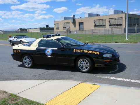 2001 Ford Crown Victoria - A black and tan state trooper car is parked on the side of the road, with a silver Corvette in the background.