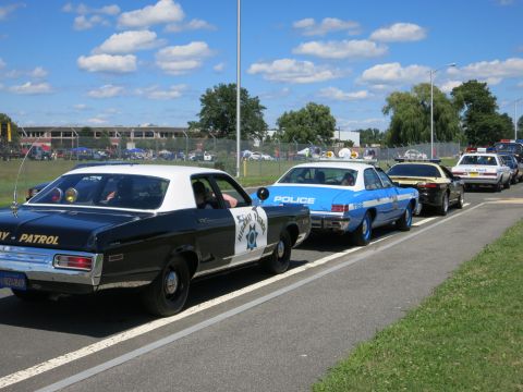 2001 Ford Crown Victoria - Several police cars, including a black and white and a blue and white model, are lined up on a paved road under a partly cloudy sky.