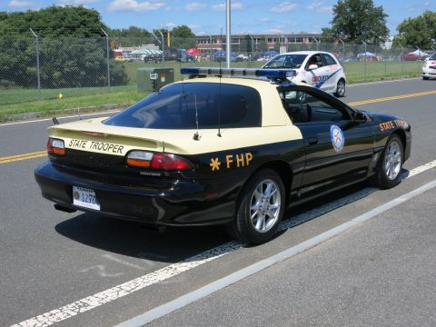2001 Ford Crown Victoria - A black and tan state trooper car is parked on the side of the road with other vehicles and tents in the background.
