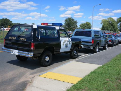 2001 Ford Crown Victoria - A line of vehicles, including a highway patrol SUV, are parked on the side of the road under a partly cloudy sky.