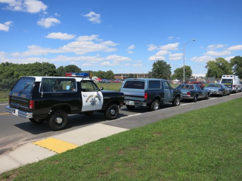 2001 Ford Crown Victoria - Several police vehicles are lined up on the side of a road under a blue sky with scattered clouds.