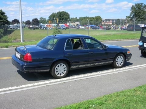 2001 Ford Crown Victoria - A dark blue 2001 Ford Crown Victoria sedan is seen from the side, driving on a road with a fairground in the background.