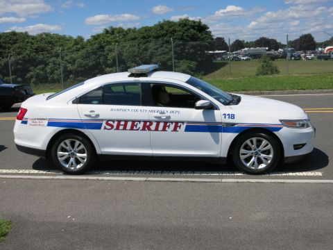 2001 Ford Crown Victoria - A white sheriff's department sedan is seen from the side, parked on a road with trees and a fence in the background.