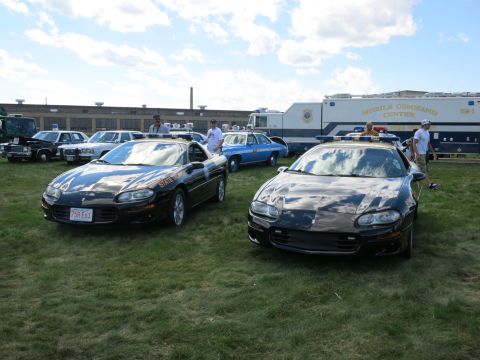 2001 Ford Crown Victoria - Two black Chevrolet Camaros, likely police vehicles, are parked on a grassy field with other vehicles and a command center truck in the bac…