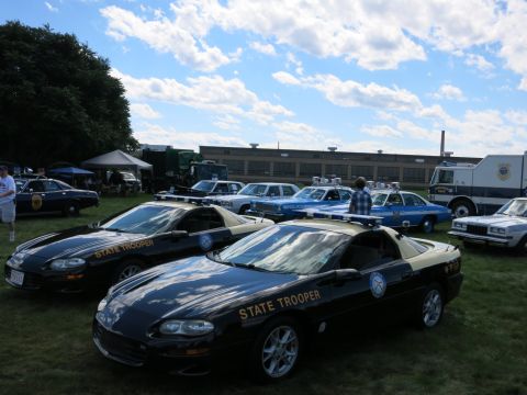 2001 Ford Crown Victoria - A collection of state trooper vehicles, including two black and tan Camaros, are displayed outdoors on a sunny day.