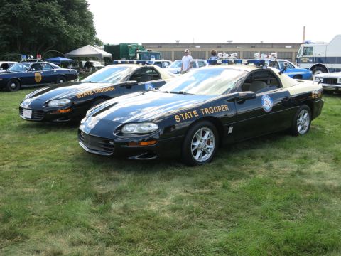 2001 Ford Crown Victoria - Two police cars, one a Chevrolet Camaro, are parked on a grassy field with other vehicles in the background.