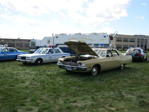 2001 Ford Crown Victoria - A tan police car with its hood open is parked on grass, with other police vehicles and a mobile command unit in the background.
