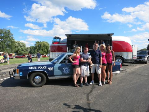 2001 Ford Crown Victoria - A group of people stand next to a blue state police car, with a large bus in the background.