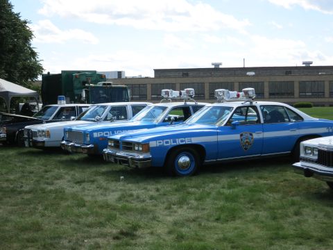 2001 Ford Crown Victoria - Several vintage police cars are lined up on a grassy field with a building in the background.