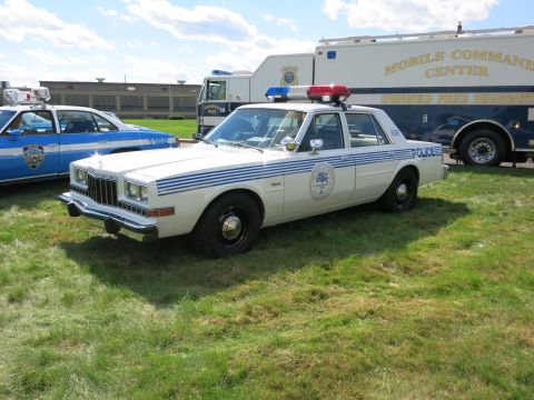 2001 Ford Crown Victoria - A white police car with blue stripes and a light bar is parked on grass next to a mobile command center.
