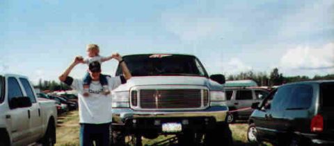 1979 Ford Bronco - A man carries a child on his shoulders in front of a large truck, with other vehicles visible in the background.