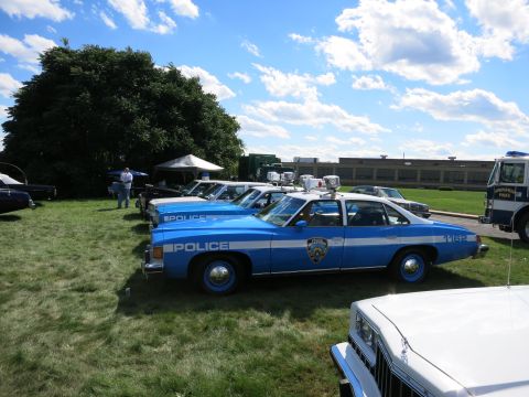2001 Ford Crown Victoria - A lineup of vintage police cruisers are displayed on a grassy field under a partly cloudy sky.
