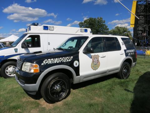 2001 Ford Crown Victoria - A black and white police SUV is parked on grass next to an ambulance under a blue sky with clouds.