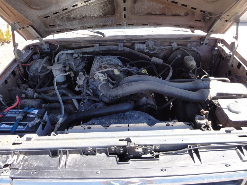 Engine bay of a 1989 Ford Bronco, showing the V8 engine, battery, and hoses. The engine compartment is dusty and appears to be in used condition