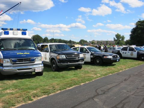 2001 Ford Crown Victoria - A line of emergency vehicles, including an ambulance and multiple police cars, are parked on a grassy area under a partly cloudy sky.