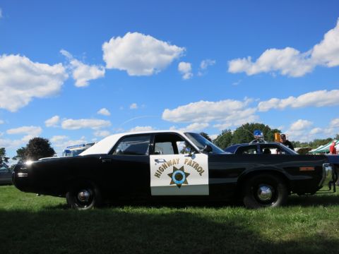 2001 Ford Crown Victoria - This vintage highway patrol car, likely from the 1970s, is parked on a grassy field under a blue sky with scattered clouds.