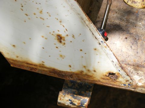 Close-up of a white metal panel with significant rust damage and a screwdriver inserted into a hole