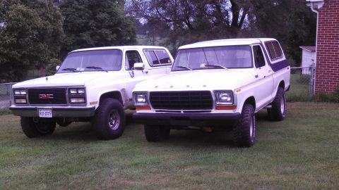 1979 Ford Bronco - A white Ford Bronco and a white GMC SUV are parked on a grassy lawn next to a brick building.